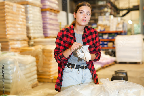 Female manager keeps records of bulk construction materials in the warehouse, writing down the data on smartphone