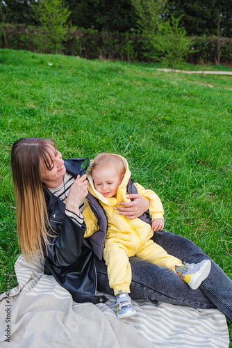 mother holds her son on her lap and sits on a blanket on a green lawn in a spring park