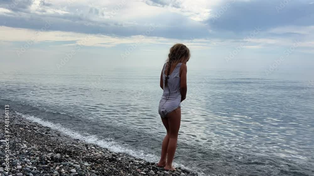 Little girl standing in the cold water of the sea on a pebble beach, cloudy sky calm