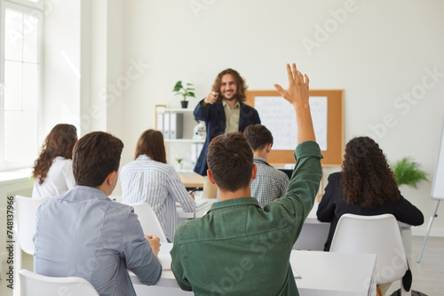 In the bustling school classroom full of students, a dedicated teenage schoolboy raises his hand, signaling active participation during a lesson. The teacher attentively guides the learning process.