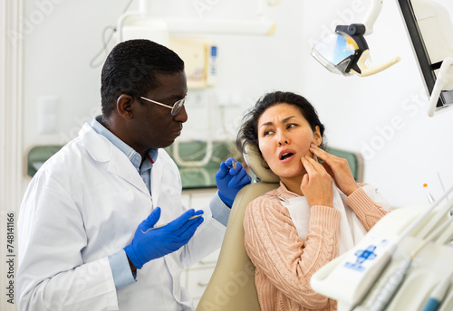 Asian woman suffering from toothache and talking about treatment with african-american man dentist.