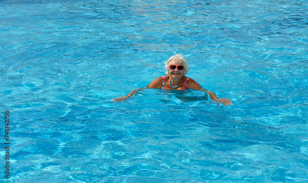 Aged woman is swimming in bright blue water of pool.