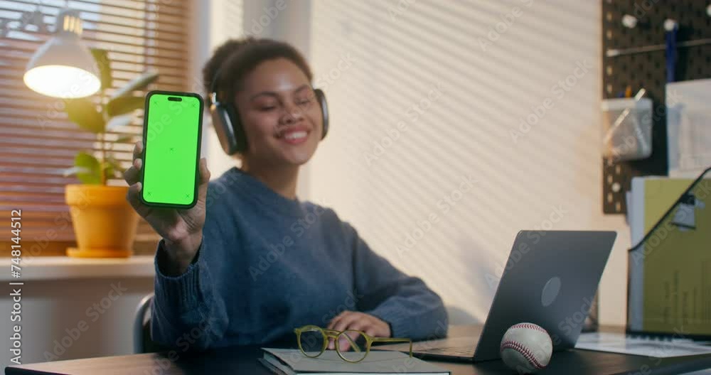 A woman in headphones smiles looking at the camera and showing a mobile phone with green screen while sitting at her desk in the office