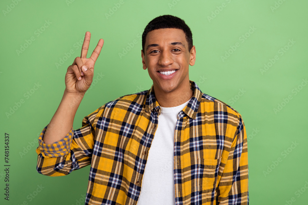 Portrait of cheerful young funny guy student wear casual plaid shirt make v sign sending greetings isolated over green color background Portrait of cheerful young funny guy student wear casual plaid shirt make v sign sending greetings isolated over green color background