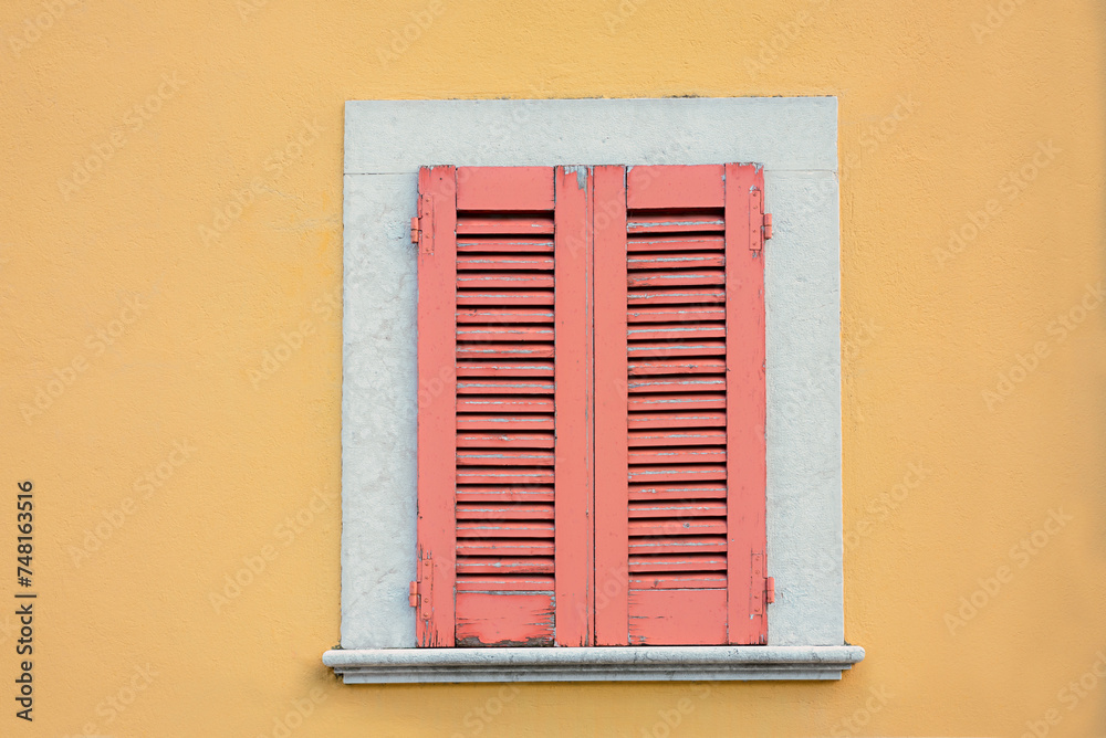 yellow house facade and window with closed red shutters, exfoliated paint