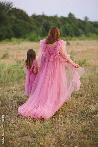 Rear view of mother and her little daughter in same tulle pink dresses holding their hands and walking on nature at summer