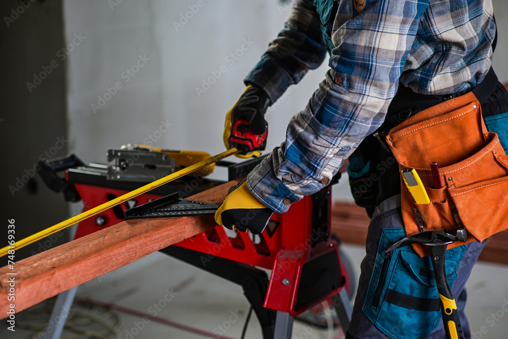 a worker in fitters and gloves with a tool belt measures wood using a tape measure and a right-angled triangle, blurred background