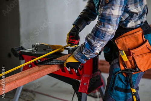 a worker in fitters and gloves with a tool belt measures wood using a tape measure and a right-angled triangle, blurred background