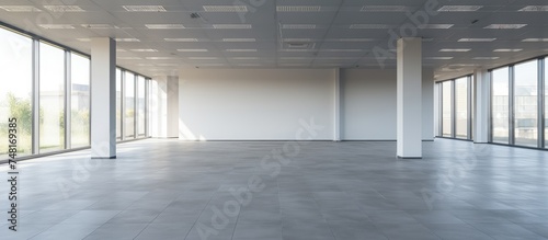 An expansive, undecorated commercial office space with a multitude of large windows letting in ample natural light. The room is vast and empty, painted in shades of gray,