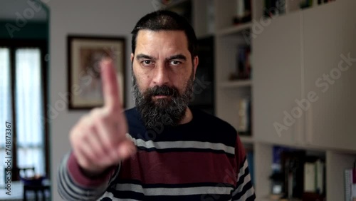 Bearded man standing with serious expression saying no with finger at home.

