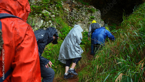 Group of tourists go to cave. Clip. Tourists enter cave in rocks on rainy day. Group of people in raincoats in rocky Mountains go to cave in summer