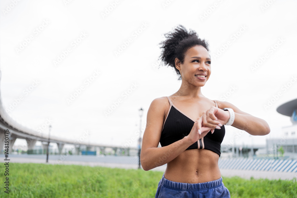 Athletic woman checking fitness tracker after a workout, with an urban landscape in the background. - obrazy, fototapety, plakaty 