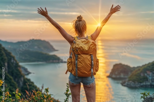 Back view of young woman with backpack standing on top of mountain and enjoying beautiful view of sea and sunset.