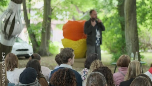 Pan left and right of male influencer talking to people sitting on chairs in park during festival in summer