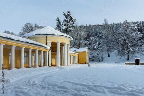 Marienbad, Czech Republic - December 4 2023: Yellow and white building of Ferdinand Spring pavilion during winter time covered with snow. Sunny evening in the park.