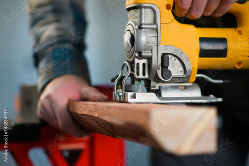 worker in overalls and wearing yellow gloves sawing wood with a reciprocating electric wood saw - close-up view and blurred background