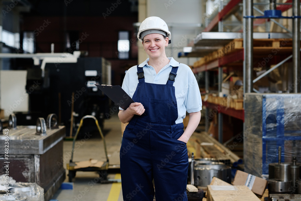 Medium long shot of cheerful young Caucasian female factory worker holding clipboard smiling at camera Medium long shot of cheerful young Caucasian female factory worker holding clipboard smiling at camera