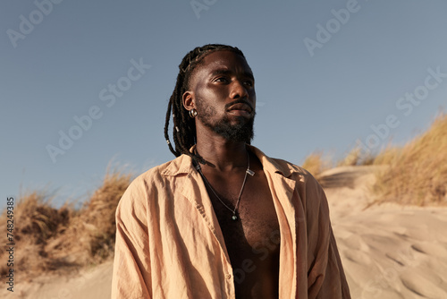 Serious young black man standing and looking away on sandy beach