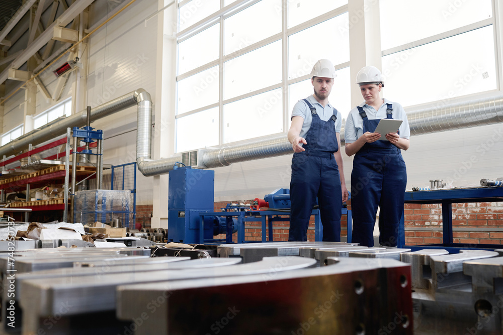 Wide shot of Caucasian man wearing hardhat demonstrating metal object produced in factory to his female colleague, copy space