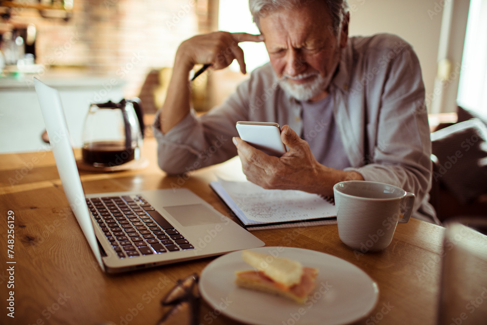 Smiling senior man using smartphone with laptop on desk at home Smiling senior man using smartphone with laptop on desk at home