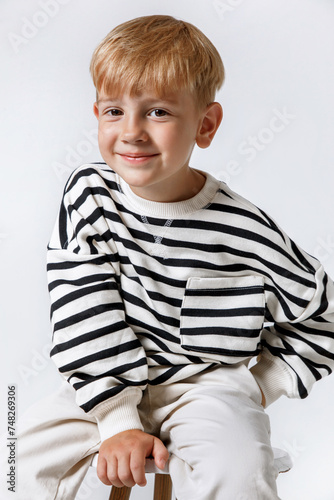 Blonde little smiling kid boy in striped sweater and white trousers sitting posing at studio
