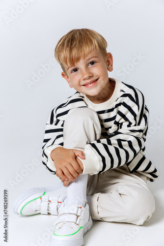 Little smiling blonde kid boy in striped sweatshirt and white trousers sitting and posing at studio