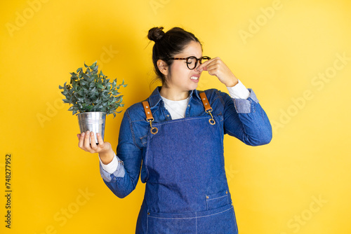 Young caucasian gardener woman holding a plant isolated on yellow background smelling something stinky and disgusting, intolerable smell, holding breath with fingers on nose