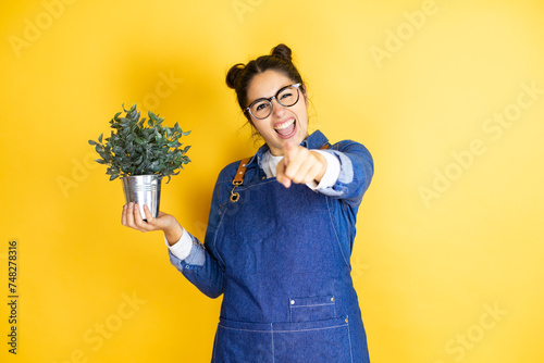 Young caucasian gardener woman holding a plant isolated on yellow background laughing at you, pointing finger to the camera with hand over body, shame expression