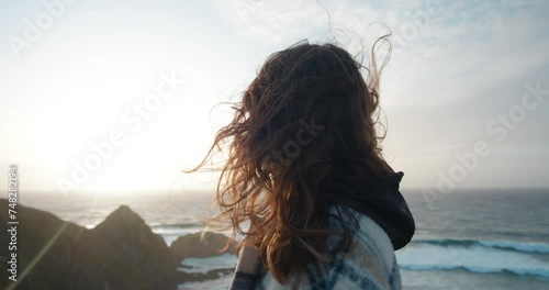 Beautiful young woman looking at the ocean or sea waves crashing into the shore. Strong ocean breeze or sea wind blow her hair. Romantic and dreamy atmosphere. Meditational concept.  photo