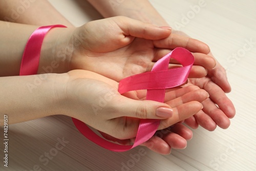 Women holding pink ribbon at white wooden table, closeup. Breast cancer awareness