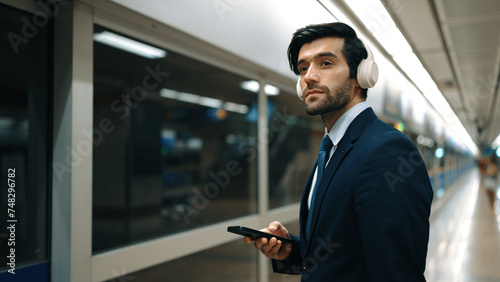 Project manager wearing headphone at train station while holding mobile phone for choosing song. Smart business man listening relaxing music while waiting for train with blurred background. Exultant.