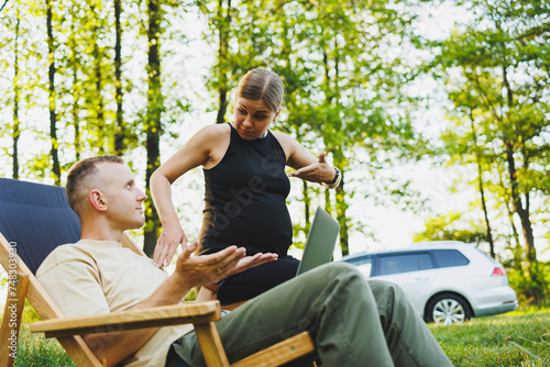 A happy couple relaxes in folding chairs near the car, a man uses a laptop for work or checking social networks, his wife treats him to a croissant. Pregnant woman with man in nature. © Дмитрий Ткачук