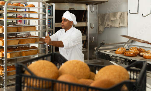 Confident baker carrying tray rack trolley with bread in industrial kitchen of bakery