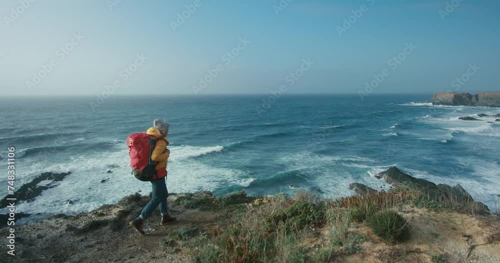 Young woman with red backpack walk to the edge of a cliff and look into cinematic ocean. Female hiker explorer. Inspiring modern outdoor lifestyle. Travel blog or advertising concept. Young woman with red backpack walk to the edge of a cliff and look into cinematic ocean. Female hiker explorer. Inspiring modern outdoor lifestyle. Travel blog or advertising concept.