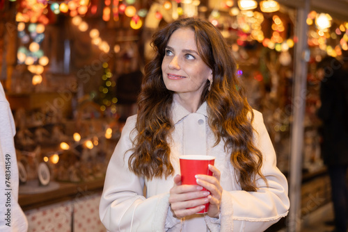 Cheerful young woman with paper cup of hot tea in hands, enjoying walking at street fair among shopping stalls offering colorful Christmas decorations