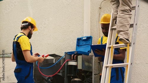 Specialists measuring freon levels in HVAC system with pressure indicators while african american coworker steps down from folding ladder after finishing checking rooftop condenser photo