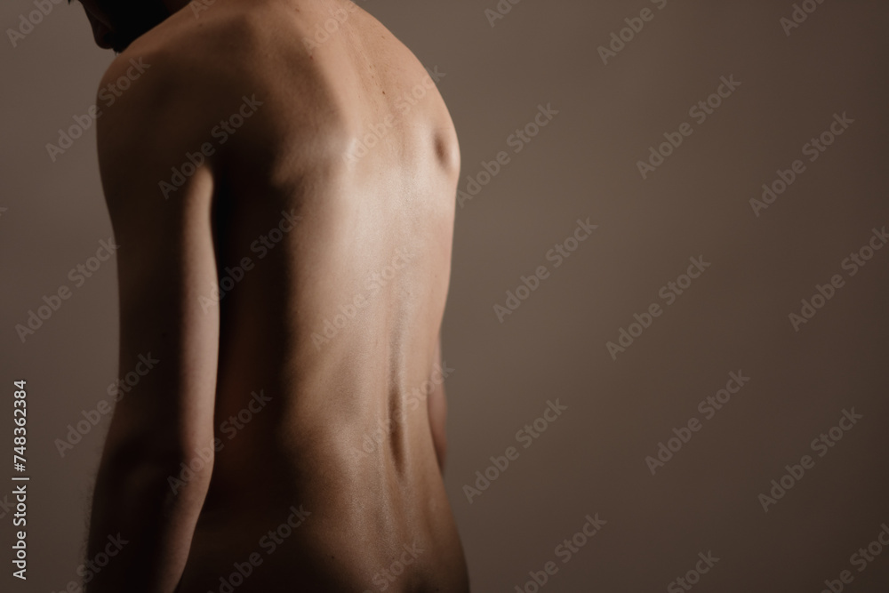 A nude man stands alone in a dimly lit studio, slouching and looking down. He has his back turned to the camera. The photo expresses themes of vulnerability, sensuality, and physical health. A nude man stands alone in a dimly lit studio, slouching and looking down. He has his back turned to the camera. The photo expresses themes of vulnerability, sensuality, and physical health.