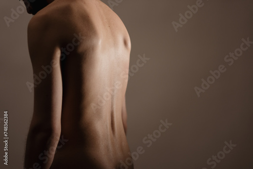 A nude man stands alone in a dimly lit studio, slouching and looking down. He has his back turned to the camera. The photo expresses themes of vulnerability, sensuality, and physical health.