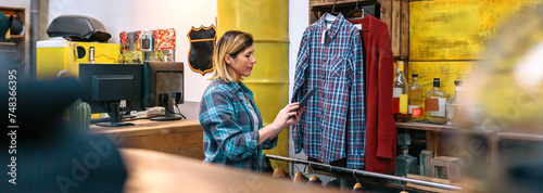 Banner of woman looking digital tablet while checking stock on second hand clothing store. Young female working on store with industrial style. Sustainable shopping and circular apparel economy. photo
