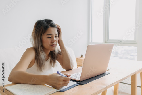 Serious freelance Asian woman is working online at home with computer laptop and taking notes in her apartment.