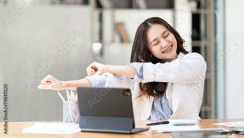 Happy relaxed young woman sitting in her office with a laptop in front of her and stretching her arms with a smile © NAMPIX