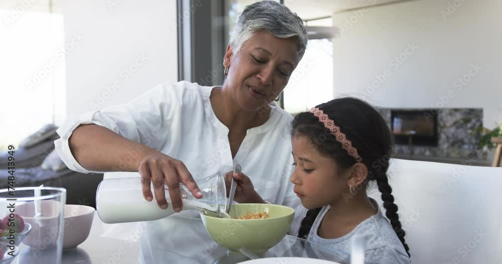 Biracial grandmother pours milk into a bowl for a young biracial granddaughter at a kitchen table Biracial grandmother pours milk into a bowl for a young biracial granddaughter at a kitchen table