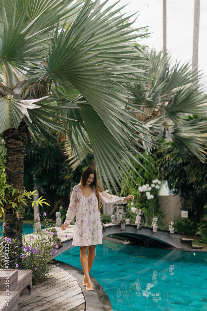Happy woman enjoying warm sunny day near infinity pool with tropical view, palm trees around, walking on the edge of the pool. Vacation on Bali island