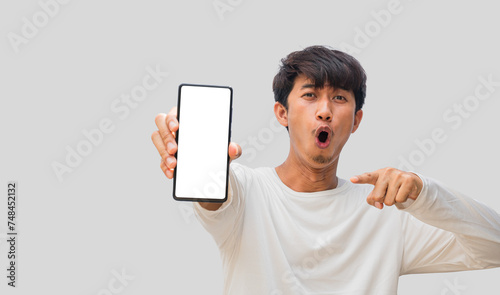 Young Asian man wearing a white t-shirt making a wow face showing mobile phone with blank white smartphone screen isolated on gray background. Guy recommending new application or mobile website