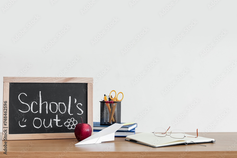 Chalkboard with text SCHOOL'S OUT, apple, paper plane and stationery on table near light wall Chalkboard with text SCHOOL'S OUT, apple, paper plane and stationery on table near light wall