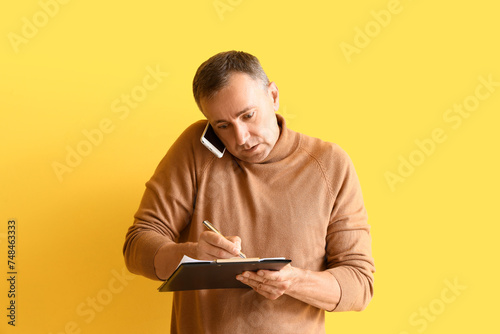 Stressed mature man with clipboard talking by mobile phone on yellow background. Deadline concept