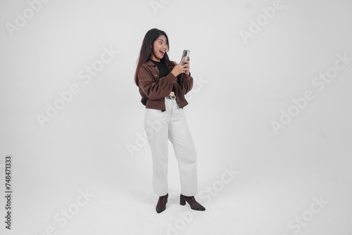 Portrait of a shocked Asian woman wearing casual outfit holding and showing her phone while her mouth wide open, standing on isolated white background