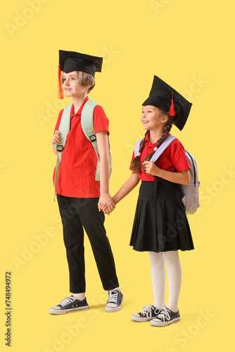 Little children in graduation hats walking on yellow background. End of school year