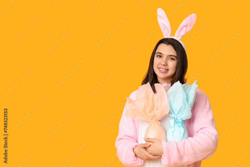 Happy young woman in bunny costume with Easter gift eggs on yellow background