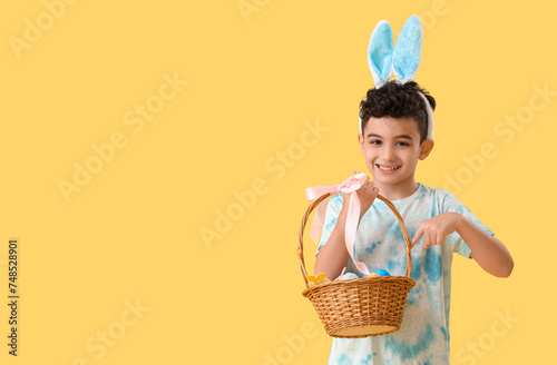 Cute little boy in bunny ears pointing at wicker basket with Easter eggs on yellow background